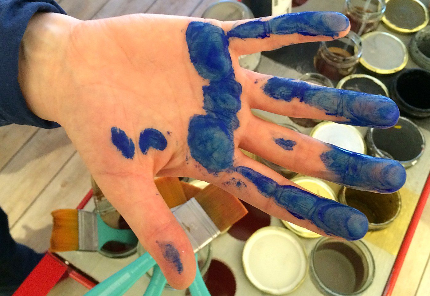 Offene Hand mit blauer Farbe an Handfläche und Fingern, im Atelier zwischen Farbgläsern und Pinseln
